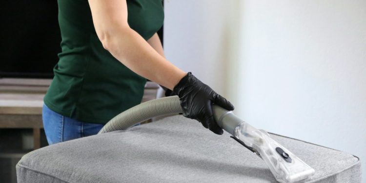 a woman in a green shirt and black gloves vacuuming a gray ottoman